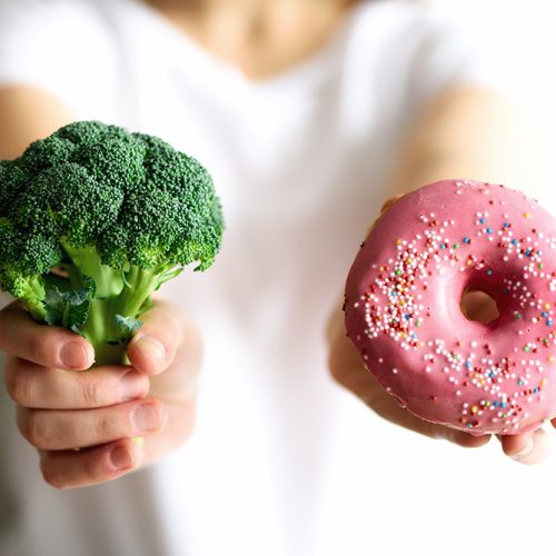 Young woman in white T-shirt choosing between broccoli or junk food, donut. Healthy clean detox eating concept. Vegetarian, vegan, raw concept. Copy space.